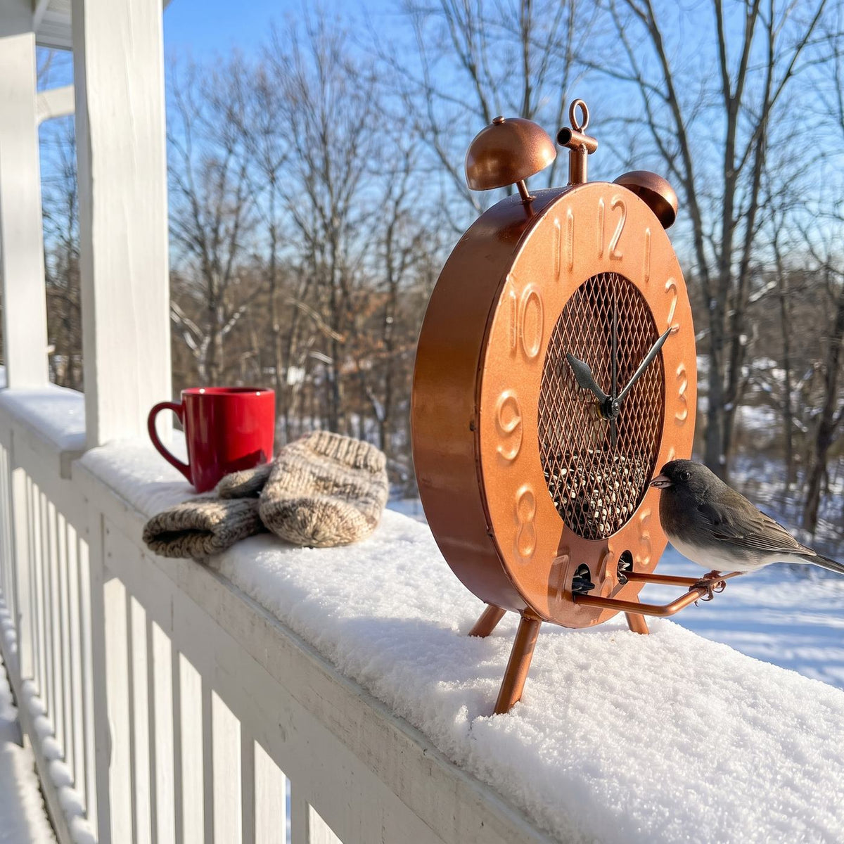 Copper clock bird feeder standing on a porch railing with a junco