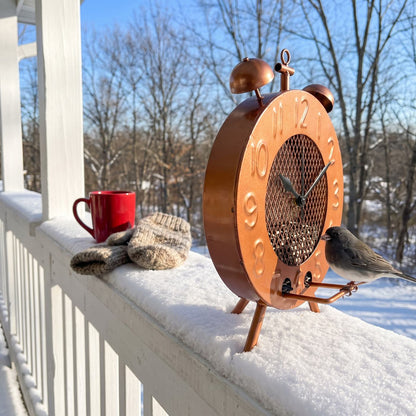 Copper clock bird feeder standing on a porch railing with a junco