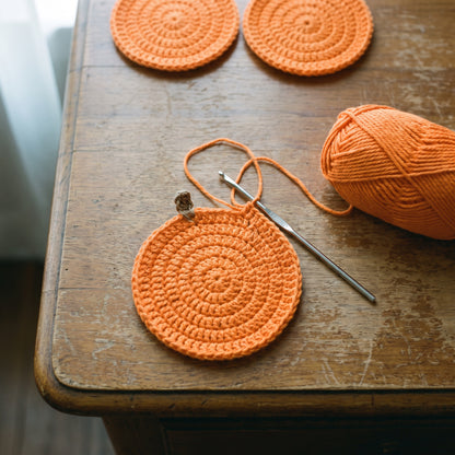 A half-finished pumpkin coaster with crochet hook and a ball of orange cotton yarn
